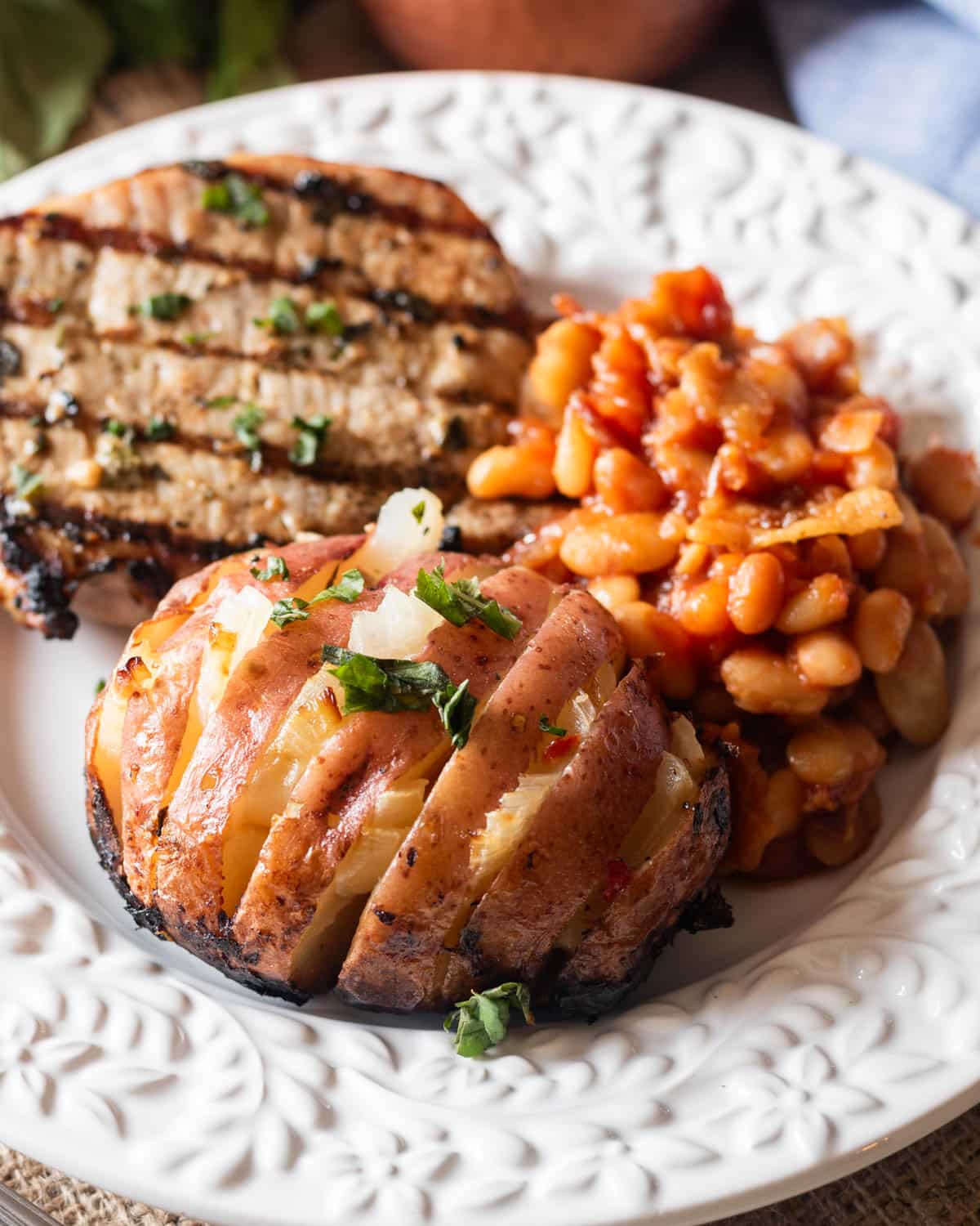 a grilled hasselback potato on a plate with beans and a pork chop