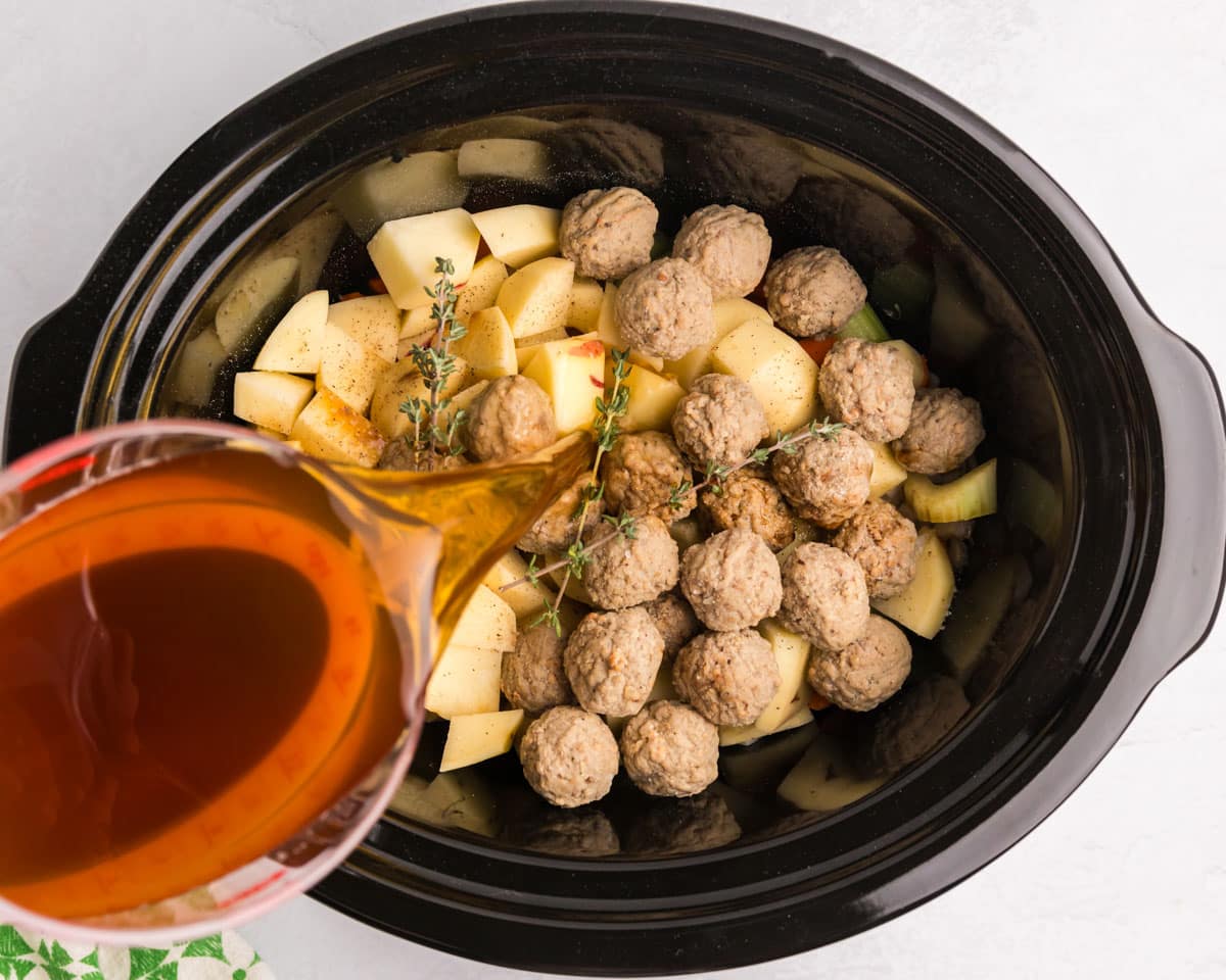 pouring beef stock over potatoes and meatballs in a slow cooker