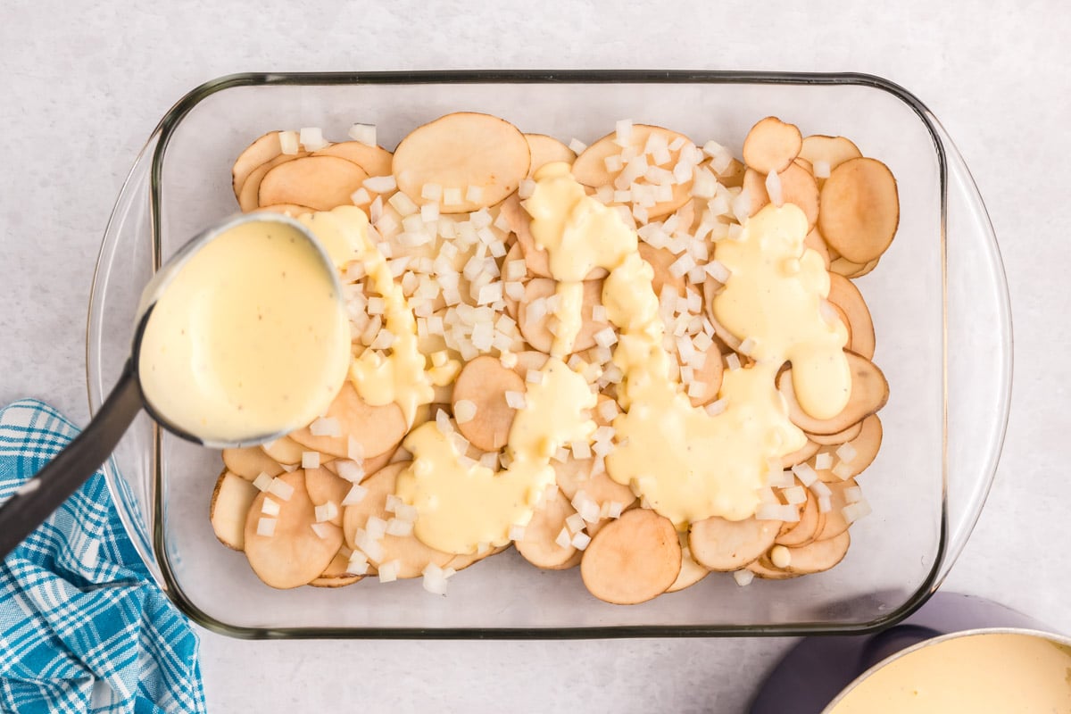 pouring cheese sauce over sliced potatoes in a casserole