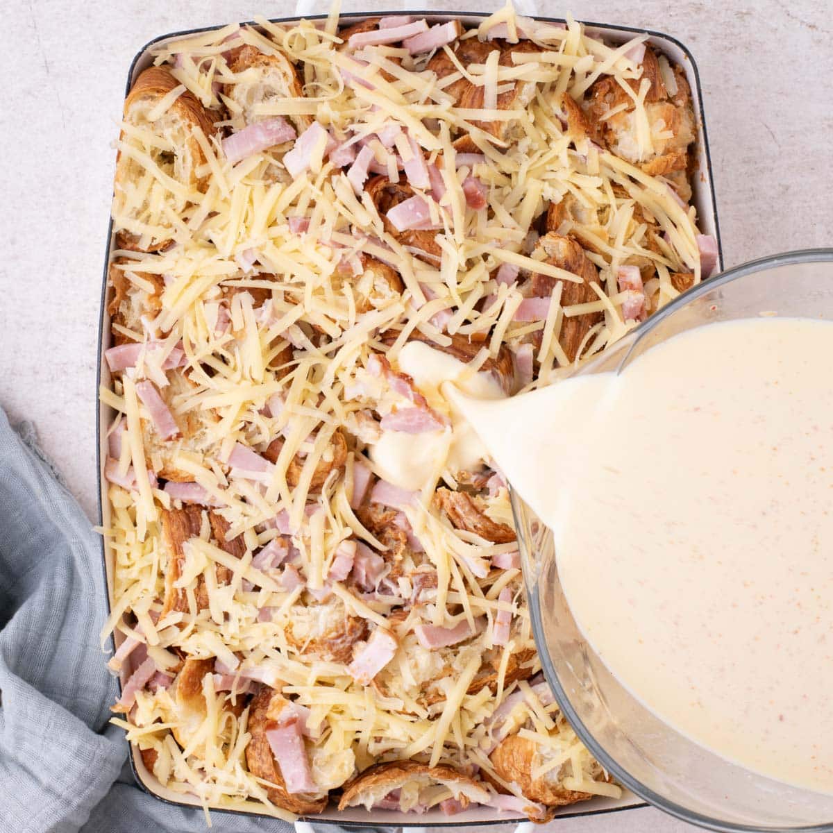 pouring egg mixture over croissants, ham and cheese in a baking dish